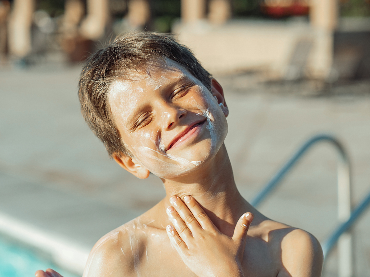 Chlapec si rozotiera po tvári opaľovací krém z prírodných surovín. Sedí blízko pri bazéne na kúpalisku.
